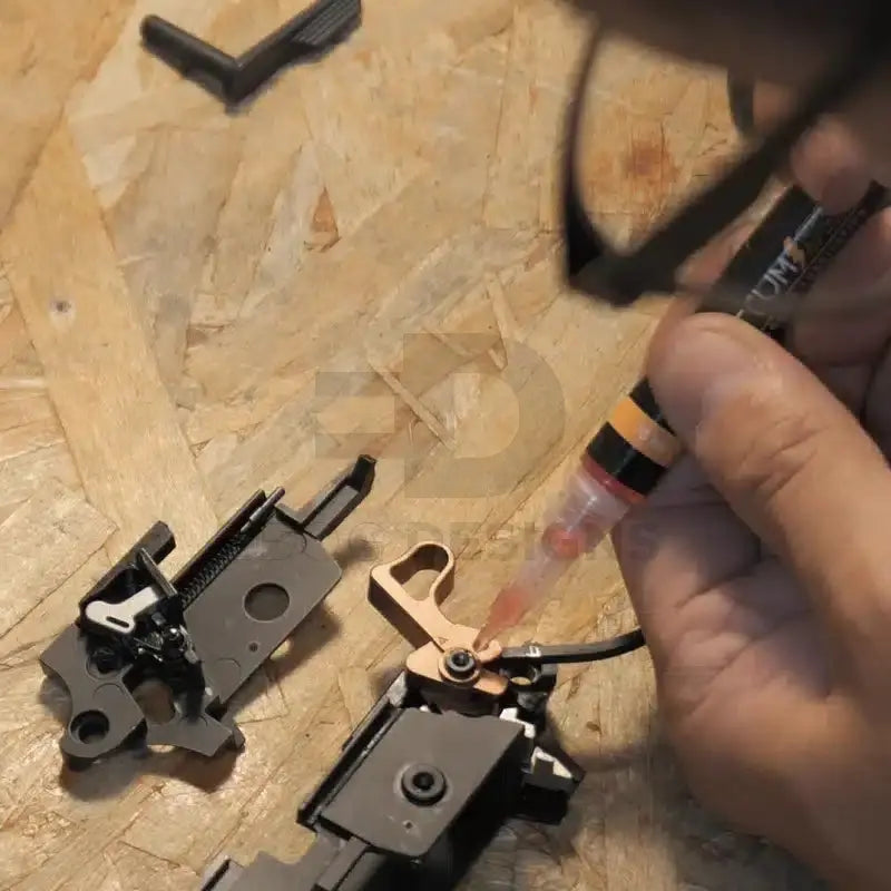 4uantum High Speed Axle and Trigger Oil applied to a disassembled airsoft trigger group with a bronze flat trigger on a workbench